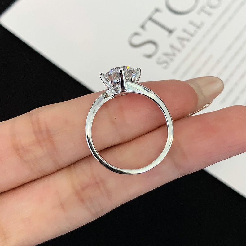 May include: A silver ring with a large, clear gemstone. The ring has a simple band and a four-prong setting that holds the stone securely. The ring is shown against a neutral background.