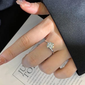 May include: A silver ring with a large, square-cut, clear gemstone. The ring is on a finger, with a dark background. The ring is a solitaire design, with a simple band.