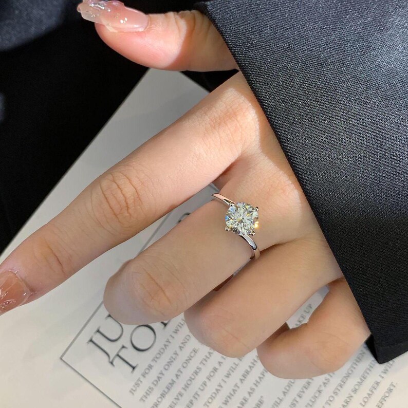May include: A silver ring with a large, square-cut, clear gemstone. The ring is on a finger, with a dark background. The ring is a solitaire design, with a simple band.