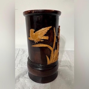 May include: A dark brown wooden vase with a carved design of a bird in flight and foliage. The vase has a cylindrical shape with a rounded base and rim. The bird and foliage are a lighter shade of brown.