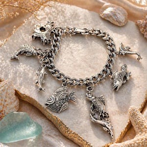 May include: A silver charm bracelet featuring various sea life charms, including fish and a seahorse. The bracelet is displayed on a textured surface with seashells and a starfish, evoking a coastal theme.