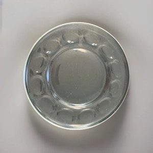 May include: Clear glass plate with a circular design. The plate has a raised rim and a series of small, rounded indentations around the edge. The center of the plate is smooth and flat. The plate is transparent and appears to be made of glass.
