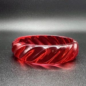 May include: A translucent red bangle bracelet featuring a continuous diagonal twisted or ribbed pattern along its entire outer surface. The circular accessory has a glossy finish and a uniform thickness, creating a distinct spiral texture throughout.
