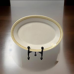 May include: An off-white oval serving platter with a gold Greek key border. The platter is displayed on a black metal stand. The design is simple and elegant, suitable for serving food.