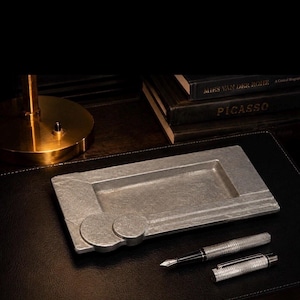 May include: A rectangular, gray metal tray with a geometric design, paired with a silver fountain pen and cap. The tray has a recessed area and circular accents. The items rest on a black leather surface, with books and a gold lamp in the background.