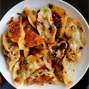 May include: A white bowl filled with a large serving of dumplings topped with a spicy sauce, sesame seeds, and green onions.