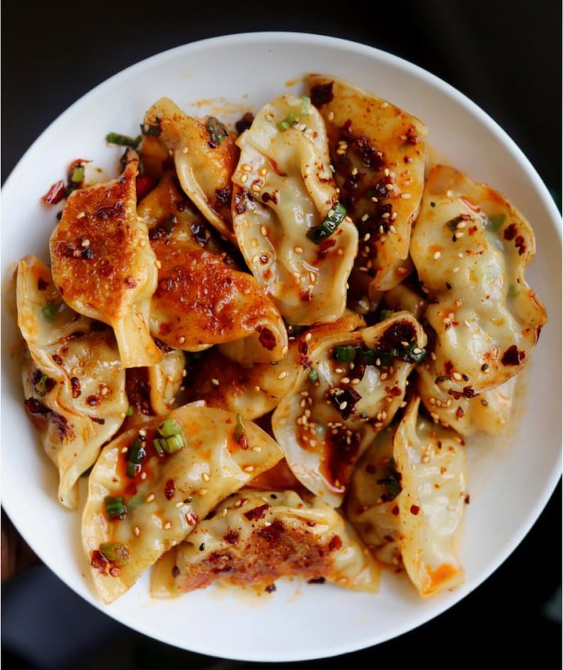 May include: A white bowl filled with a large serving of dumplings topped with a spicy sauce, sesame seeds, and green onions.