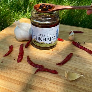 May include: A glass jar of Laza De Bukhara Premium Quality Chili Oil, with a wooden spoon scooping out some of the oil and chili flakes. The jar is on a wooden cutting board with red chili peppers and garlic cloves.