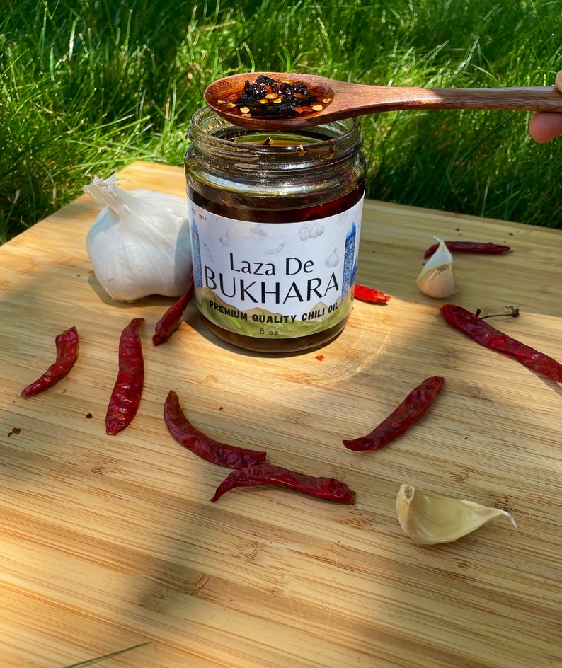 May include: A glass jar of Laza De Bukhara Premium Quality Chili Oil, with a wooden spoon scooping out some of the oil and chili flakes. The jar is on a wooden cutting board with red chili peppers and garlic cloves.