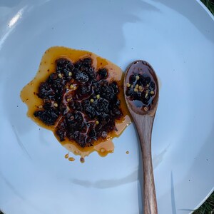 May include: A white plate with a small wooden spoon and a pile of dark brown chili flakes in a red oil sauce.