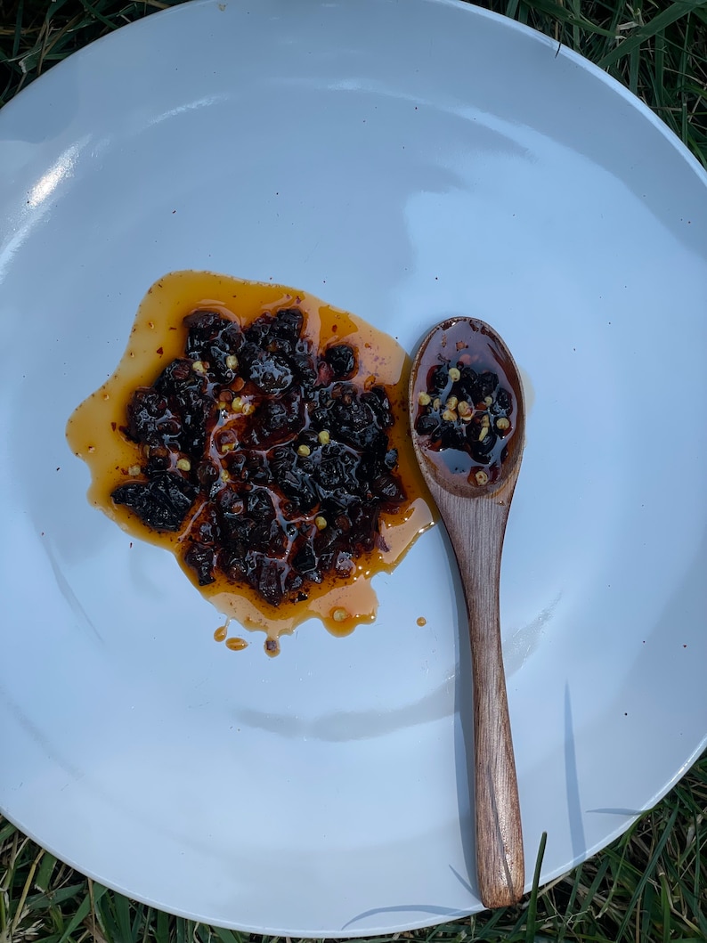 May include: A white plate with a small wooden spoon and a pile of dark brown chili flakes in a red oil sauce.