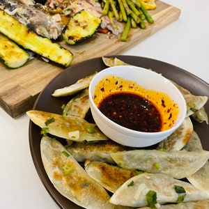 May include: A plate of pan-fried dumplings with a side of spicy red chili sauce. The dumplings are golden brown and have a crispy texture. The sauce is a vibrant red color and has a spicy flavor.