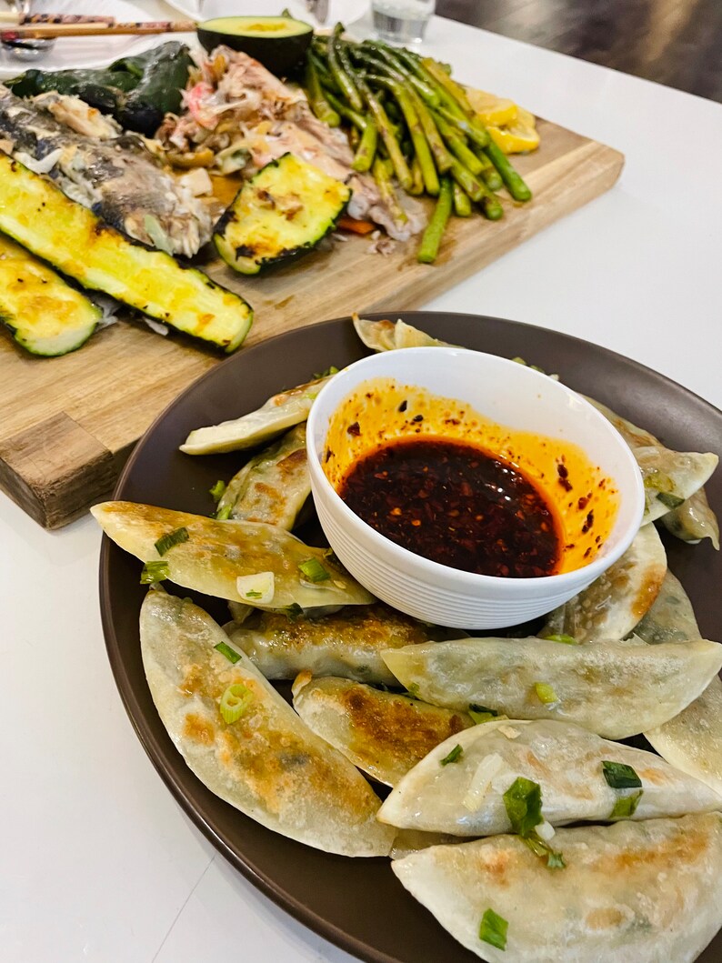 May include: A plate of pan-fried dumplings with a side of spicy red chili sauce. The dumplings are golden brown and have a crispy texture. The sauce is a vibrant red color and has a spicy flavor.