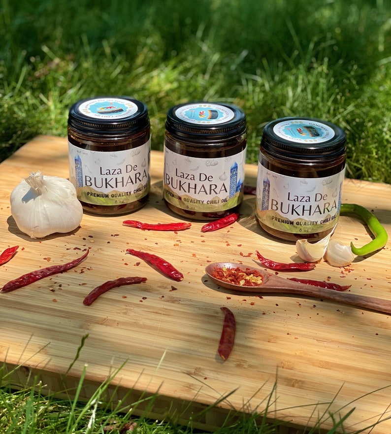 May include: Three glass jars of Laza De Bukhara premium quality chili oil on a wooden surface with red chili peppers and garlic cloves.
