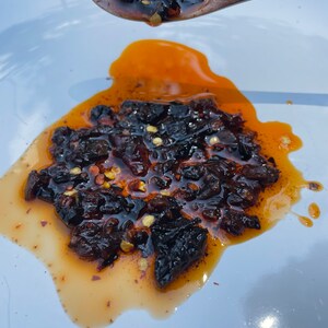 May include: A white plate with a small pile of dark brown chili peppers in a red oil sauce. The chili peppers are scattered on the plate and some are still on a wooden spoon.