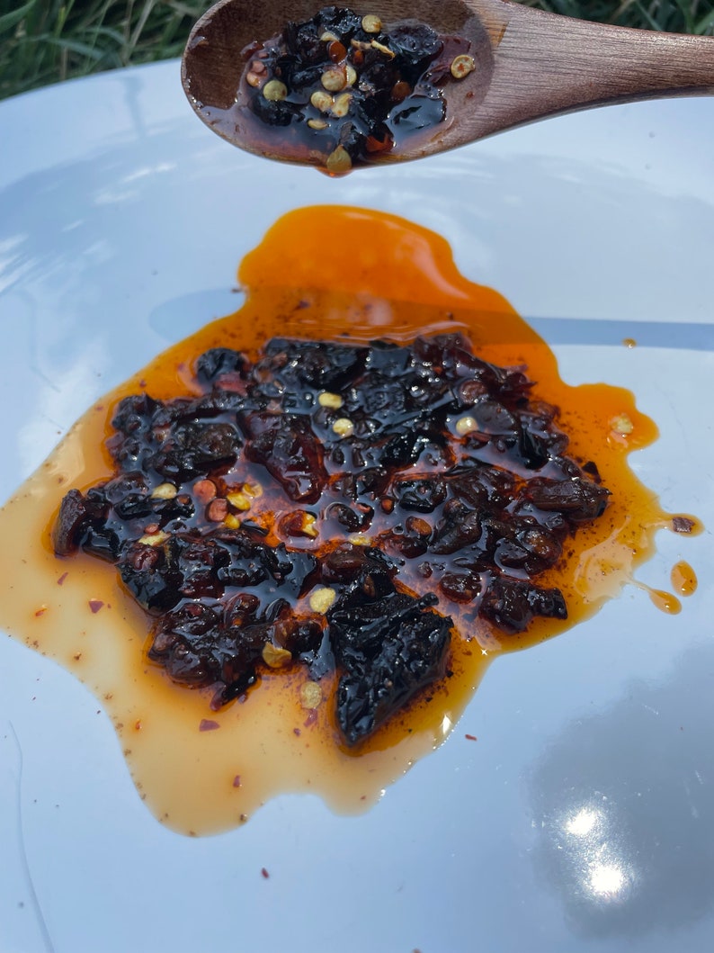 May include: A white plate with a small pile of dark brown chili peppers in a red oil sauce. The chili peppers are scattered on the plate and some are still on a wooden spoon.