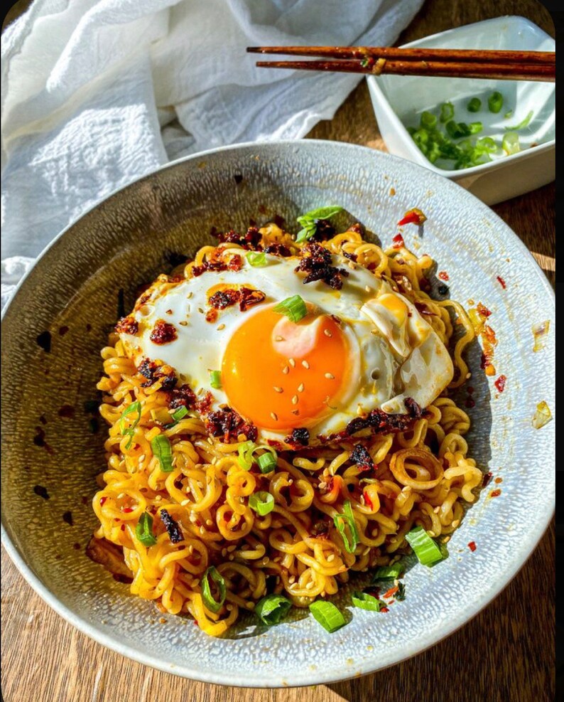 May include: A bowl of ramen noodles topped with a fried egg, green onions, and chili flakes.