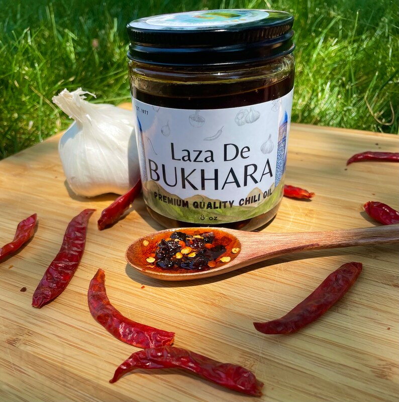 May include: A glass jar of Laza De Bukhara Premium Quality Chili Oil with a white label and a spoon with a dollop of chili oil. The jar is sitting on a wooden surface with dried red chili peppers.