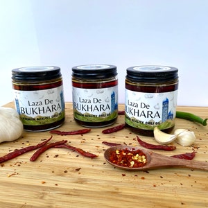 May include: Three glass jars of Laza De Bukhara premium quality chili oil with black lids. The jars are on a wooden surface with red chili peppers, garlic cloves, and a small wooden spoon with red chili flakes.