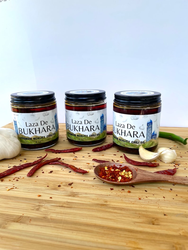 May include: Three glass jars of Laza De Bukhara premium quality chili oil with black lids. The jars are on a wooden surface with red chili peppers, garlic cloves, and a small wooden spoon with red chili flakes.