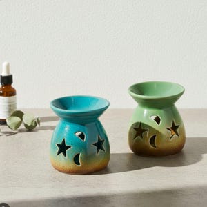 Puede incluir: Dos quemadores de aceite de cerámica. Uno es turquesa con recortes de estrellas y medias lunas, el otro es verde con recortes de estrellas y medias lunas. Ambos tienen una base marrón y una parte superior redondeada para el aceite. Una botella de aceite está en el fondo.