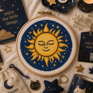 May include: A circular wooden embroidery hoop containing a cross-stitch pattern of a yellow sun with a smiling face against a dark blue background filled with small white stars and gold plus signs, centered among various celestial-themed items.