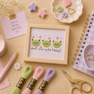 May include: A framed cross-stitch featuring three green frog faces with the text "don't worry, be happy!". The image includes embroidery floss in green, pink, and purple, along with a notepad, scissors, and decorative elements.