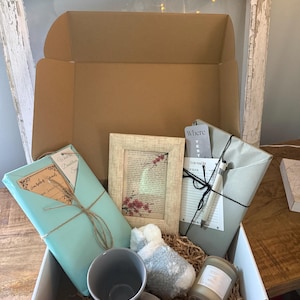 Op de afbeelding: Een open cadeauverpakking met diverse items, waaronder een ingelijste foto met bloemmotieven, een grijze mok, een kaars en verpakte pakketten. De doos toont de inhoud tegen een neutrale achtergrond.