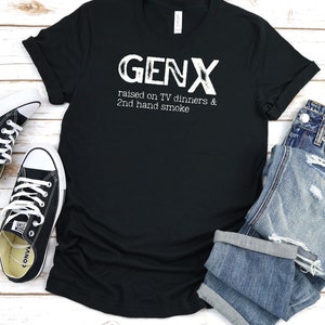 May include: Black t-shirt with white text that reads "GENX raised on TV dinners & 2nd hand smoke". The shirt is laid flat on a white wooden surface with a pair of black sneakers and a pair of blue jeans.