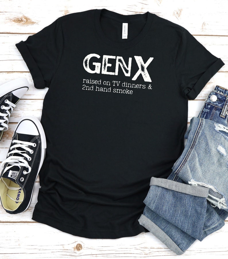 May include: Black t-shirt with white text that reads "GENX raised on TV dinners & 2nd hand smoke". The shirt is laid flat on a white wooden surface with a pair of black sneakers and a pair of blue jeans.