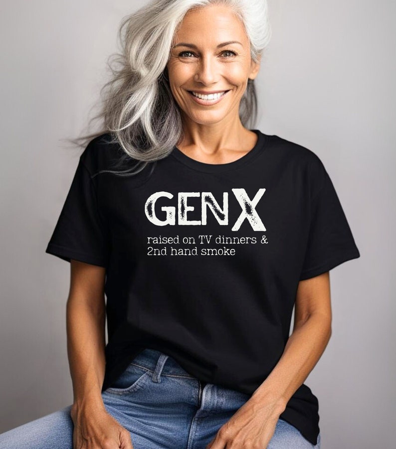 May include: A black t-shirt with white text that reads "GENX raised on TV dinners & 2nd hand smoke". A woman is wearing the shirt and smiling at the camera.