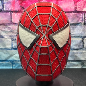 May include: A red Spider-Man mask with a web pattern and silver detailing. The mask has white mesh eye coverings and is displayed on a stand. The background is a brick wall.