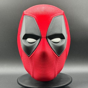 May include: A red and black Deadpool mask with a textured surface. The mask has a white eye mesh and a black eye outline.