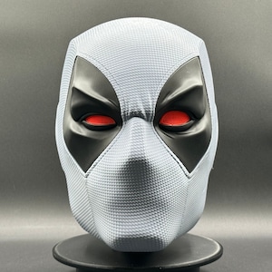 Może przedstawiać: Biała maska Deadpoola z czarnymi otworami na oczy i czerwonymi oczami. Maska ma fakturowaną powierzchnię.