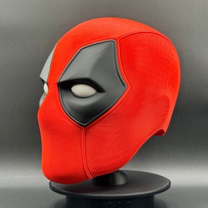 K&ouml;nnte beinhalten: Eine rote und schwarze Deadpool-Maske mit einem wei&szlig;en Augennetz. Die Maske steht auf einem schwarzen St&auml;nder.