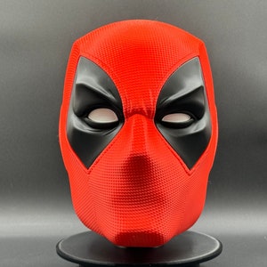 Puede incluir: Una m&aacute;scara de Deadpool roja y negra con una superficie texturizada. La m&aacute;scara tiene ojos blancos y una boca negra.