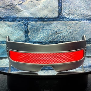 May include: A silver-colored visor with a bright red, grid-patterned lens. The visor has a curved design and is displayed against a blue brick wall background. The visor is a cosplay accessory.