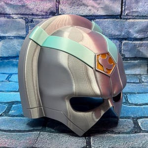 May include: A silver and iridescent helmet with a teal band and a gold emblem. The helmet has a visor and is made of a metallic material. The emblem features a bird-like design.
