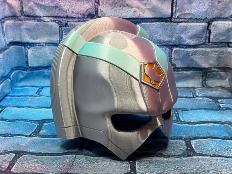 May include: A silver and iridescent helmet with a teal band and a gold emblem. The helmet has a visor and is made of a metallic material. The emblem features a bird-like design.