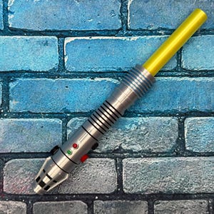 May include: A silver and black toy lightsaber with a yellow blade. The handle has a series of black lines and silver rings. It features red and green buttons. The lightsaber is set against a blue brick background.