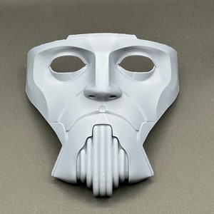 May include: A white plastic mask with a human-like face, featuring two eye holes and a detailed mouth with a series of vertical ridges.