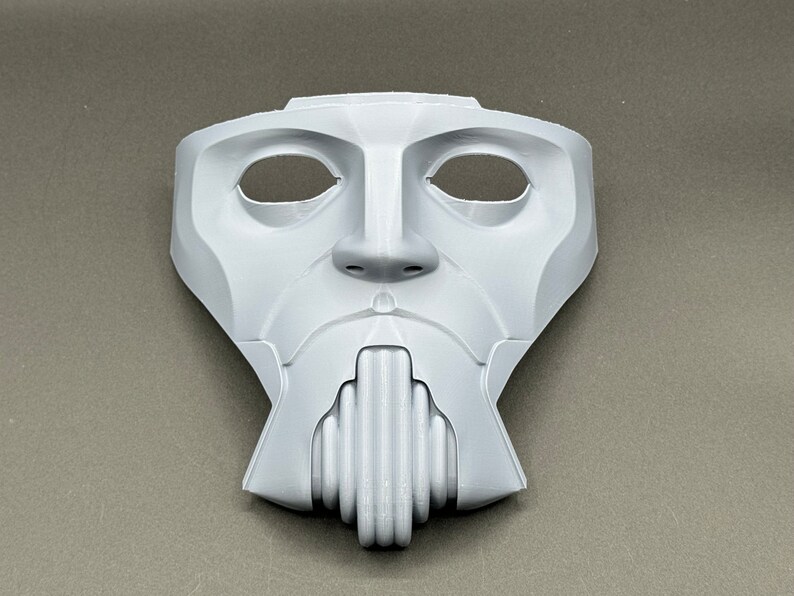 May include: A white plastic mask with a human-like face, featuring two eye holes and a detailed mouth with a series of vertical ridges.