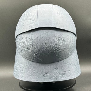 May include: A white helmet with a textured surface. The helmet has a raised ridge on the top and a visor.