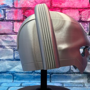 May include: A silver-colored helmet with a vertical, ridged accent on top. The helmet has a smooth, metallic finish and a visor-like opening for the eyes. It is displayed on a black stand against a brick wall.