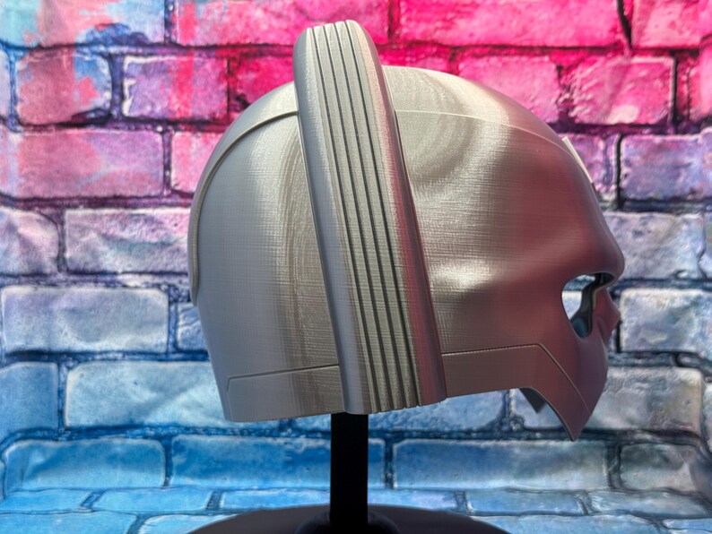 May include: A silver-colored helmet with a vertical, ridged accent on top. The helmet has a smooth, metallic finish and a visor-like opening for the eyes. It is displayed on a black stand against a brick wall.