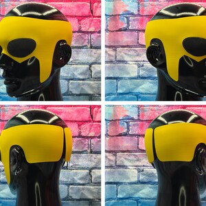 May include: A bright yellow superhero-style mask featuring a smooth, contoured design that covers the forehead and temples, with large oval cutouts for the eyes. The mask is displayed on a glossy black mannequin head from multiple angles.