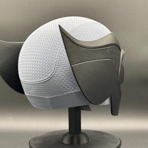 May include: A black and gray 3D printed helmet with a textured surface and a pointed ear design. The helmet is on a black stand.