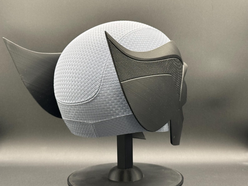May include: A black and gray 3D printed helmet with a textured surface and a pointed ear design. The helmet is on a black stand.