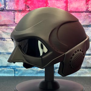 May include: A matte black helmet with a visor and a face mask. The helmet is displayed on a black stand against a brick wall background. The visor is reflective, and the face mask has a circular design.