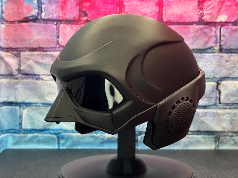 May include: A matte black helmet with a visor and a face mask. The helmet is displayed on a black stand against a brick wall background. The visor is reflective, and the face mask has a circular design.